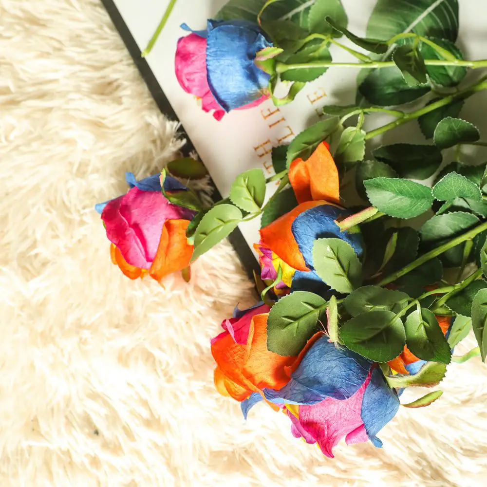 Un ramo de rosas con pétalos teñidos de varios colores, como azul, rosa y naranja. Las rosas están dispuestas con hojas verdes y colocadas sobre una superficie texturizada que parece ser una tela blanca y esponjosa.