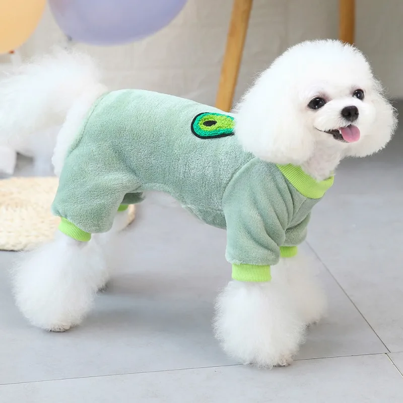 Un perro pequeño, probablemente un Bichon Frise, que lleva un suéter verde para perros con un lindo diseño verde y amarillo en la espalda. El perro tiene una bata blanca esponjosa y está parado sobre un suelo de baldosas.