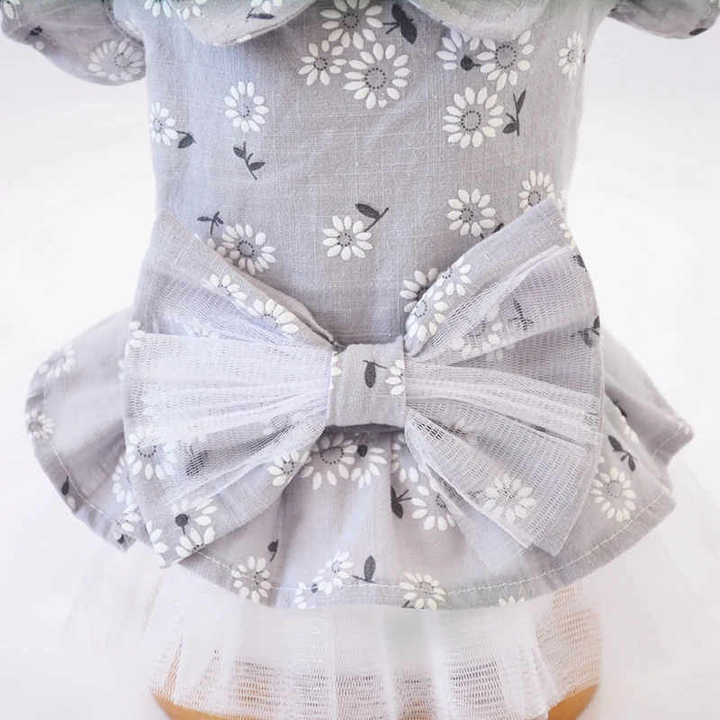 Una muñeca que lleva un vestido con estampado de flores. El vestido tiene un fondo gris claro con flores blancas y hojas verdes. Presenta un lazo en la espalda, que es un elemento de diseño común en la ropa infantil.