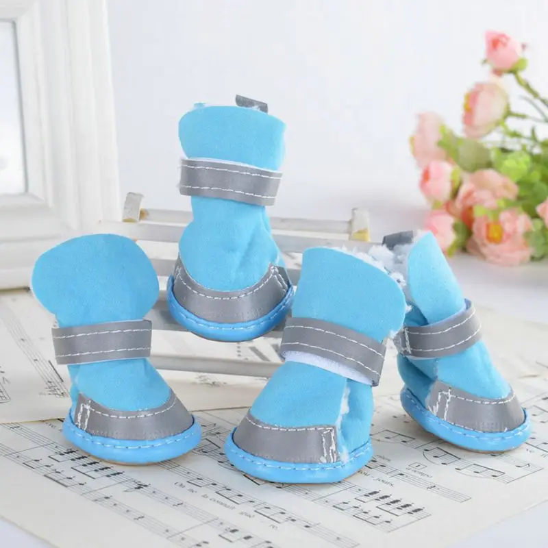 Un par de pantuflas azules con una tira gris en la parte superior. Están colocados sobre lo que parece ser una partitura y hay un jarrón con flores al fondo. Las pantuflas tienen una textura afelpada y parecen ser para niños o adultos pequeños.