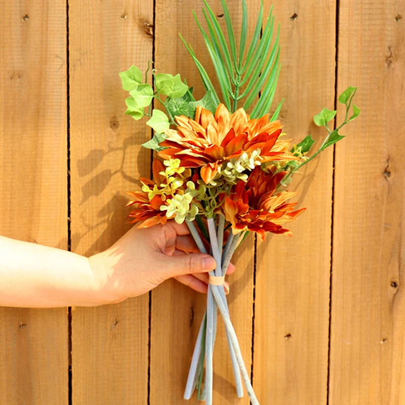 Una mano sosteniendo un ramo de flores sobre un fondo de madera. El ramo consta de una variedad de flores, incluidas lo que parecen ser flores de color naranja y amarillo, posiblemente lirios o gladiolos, y algo de vegetación.