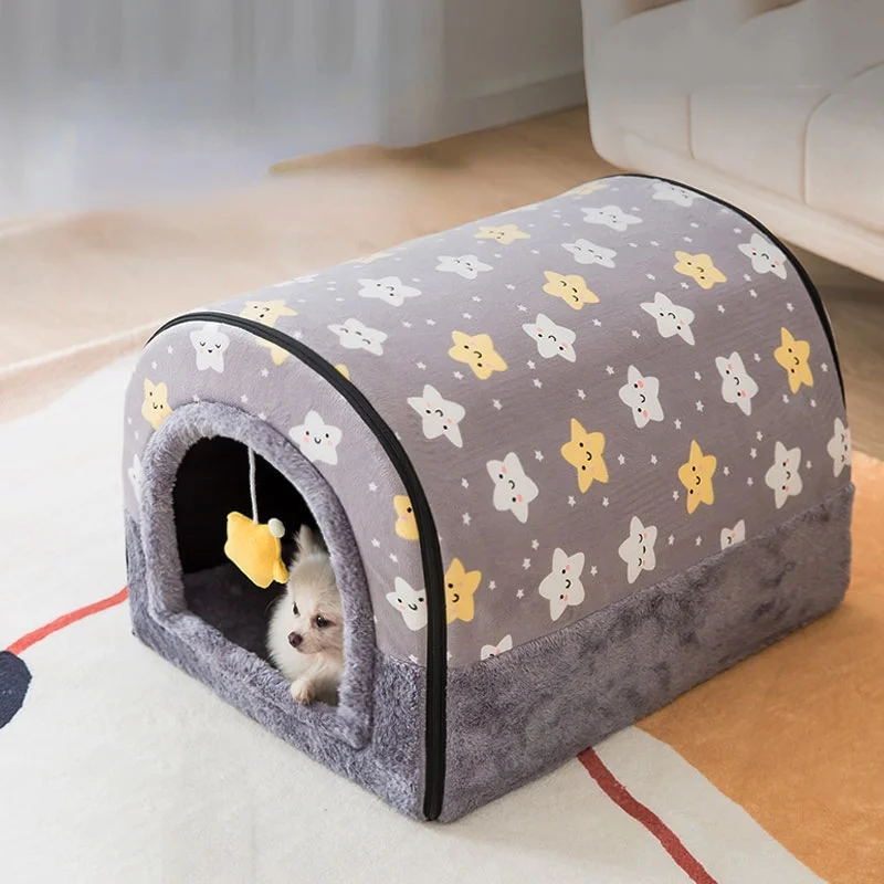 Un pequeño perro blanco sentado dentro de una cama para mascotas. La cama tiene una combinación de colores gris y blanco con un patrón de estrellas y lo que parecen ser caras de animales caricaturescas. La cama está diseñada para proporcionar un espacio acogedor y confortable para que el perro descanse o duerma.