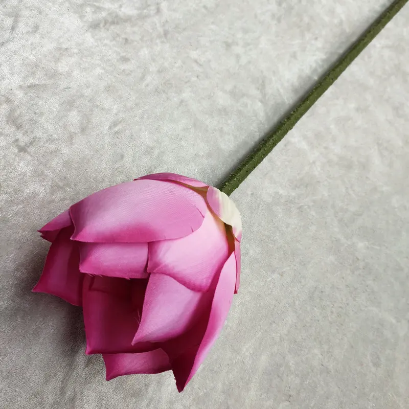 Una sola rosa rosa con un tallo verde. La rosa parece fresca y se coloca sobre un fondo texturizado que parece una superficie de tela o papel. La rosa está en plena floración, con sus pétalos completamente desplegados, dejando al descubierto los estambres en el centro.