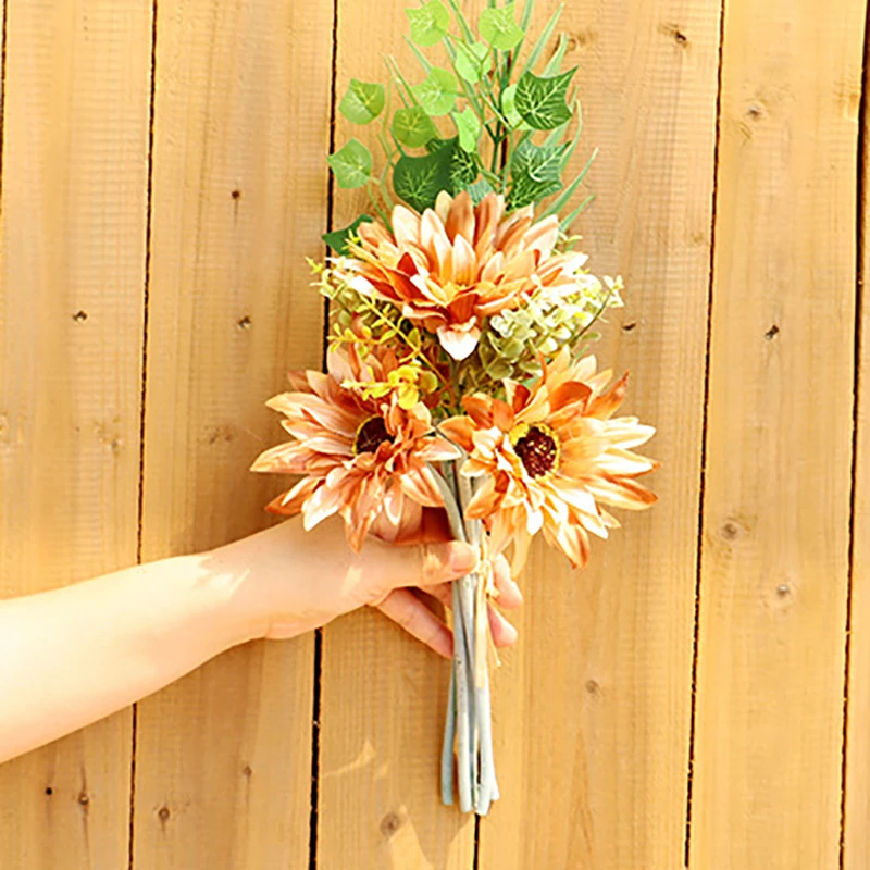 Una mano sosteniendo un ramo de flores sobre un fondo de madera. El ramo consta de una variedad de flores, incluidos lo que parecen ser girasoles y posiblemente algo de vegetación o follaje.