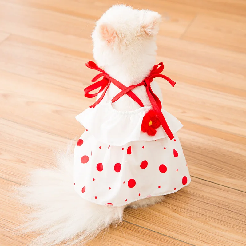 Un pequeño peluche blanco, que parece un gato, vestido con un vestido blanco con lunares rojos. El vestido tiene una cinta roja atada al cuello del peluche. El animal de peluche está sentado sobre un suelo de madera.