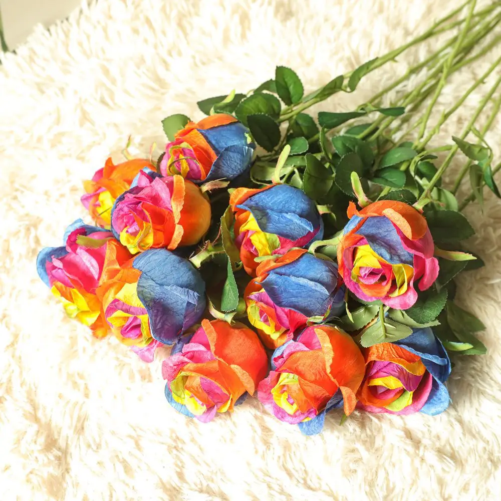 Un ramo de rosas de colores con varios tonos de naranja, rosa, azul y amarillo. Las rosas parecen frescas y están dispuestas con hojas y tallos verdes. El ramo se coloca sobre una superficie blanca y esponjosa que puede ser un cojín o una tela suave.