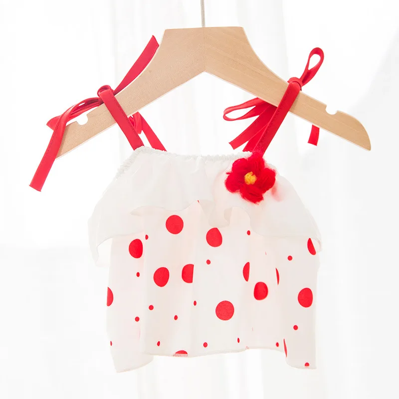 Un vestido infantil colgado de una percha de madera. El vestido tiene un fondo blanco con lunares rojos y presenta un detalle de flor roja en la parte delantera. La percha tiene cintas rojas atadas alrededor, añadiendo un toque decorativo.