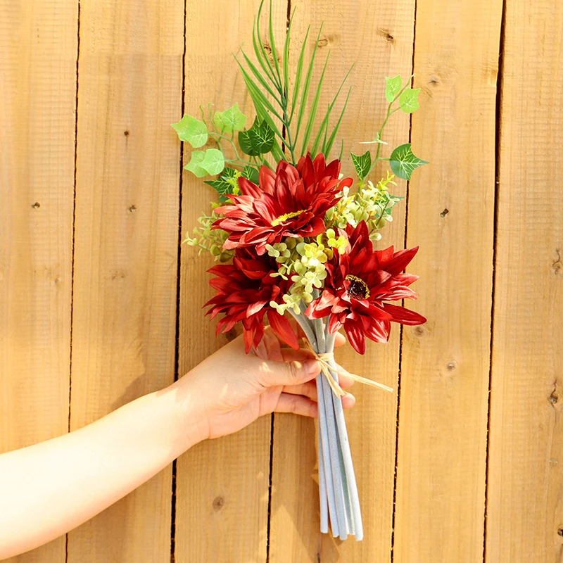 Una mano sosteniendo un ramo de flores sobre un fondo de madera. El ramo consta de flores rojas, posiblemente rosas y algo de vegetación, incluido lo que parece ser aliento de bebé y posiblemente algunos helechos u otro follaje.