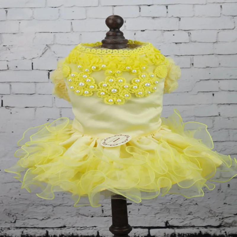 Un vestido amarillo exhibido sobre un maniquí. El vestido presenta una falda con volantes y un corpiño adornado con lo que parece ser encaje y posiblemente algunos adornos. El maniquí está colocado contra una pared de ladrillos, lo que proporciona un fondo que contrasta con el amarillo brillante del vestido.