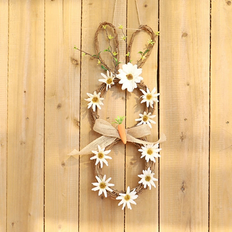 Una corona decorativa elaborada con materiales naturales. Presenta una base circular hecha de ramitas o ramas, adornada con flores blancas que parecen margaritas y una zanahoria en el centro. La corona se ata con un lazo hecho de una tela o cinta de color claro.