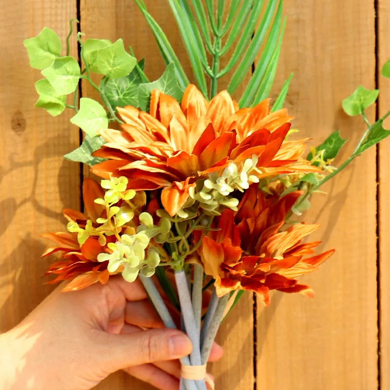 Un ramo de flores sostenido por una persona. El ramo consta de una variedad de flores con diferentes colores y texturas, incluidos tonos de naranja, amarillo y verde. También hay algunas hojas verdes y posiblemente algo de follaje o ramas mezcladas con las flores.