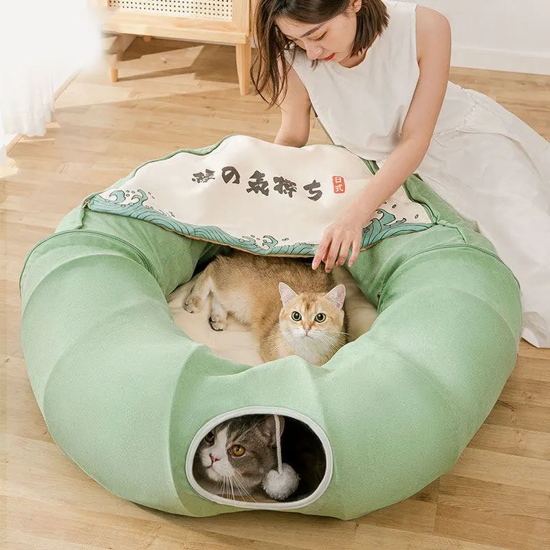 Una persona sentada en el suelo con una cama para gatos. Hay dos gatos en la cama; uno está acostado y el otro se asoma desde la entrada de la cama. La cama para gatos tiene una funda con un patrón y lo que parecen ser caracteres japoneses.