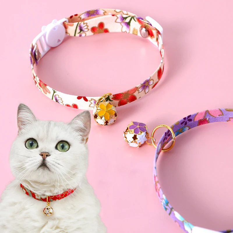 Un gato blanco con expresión de sorpresa o curiosidad. El gato lleva un collar rojo con una campana dorada. Al fondo, hay dos collares para gatos adicionales con motivos florales, uno en tono rosado y otro en tono violeta.