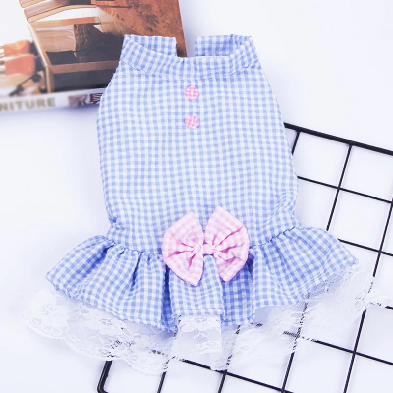 Vestido infantil con estampado de cuadros azules y blancos. El vestido tiene cuello, manga corta y un lazo en la parte delantera. Parece ser un vestido casual, posiblemente de verano, y se muestra sobre un fondo blanco con un patrón de cuadrícula.