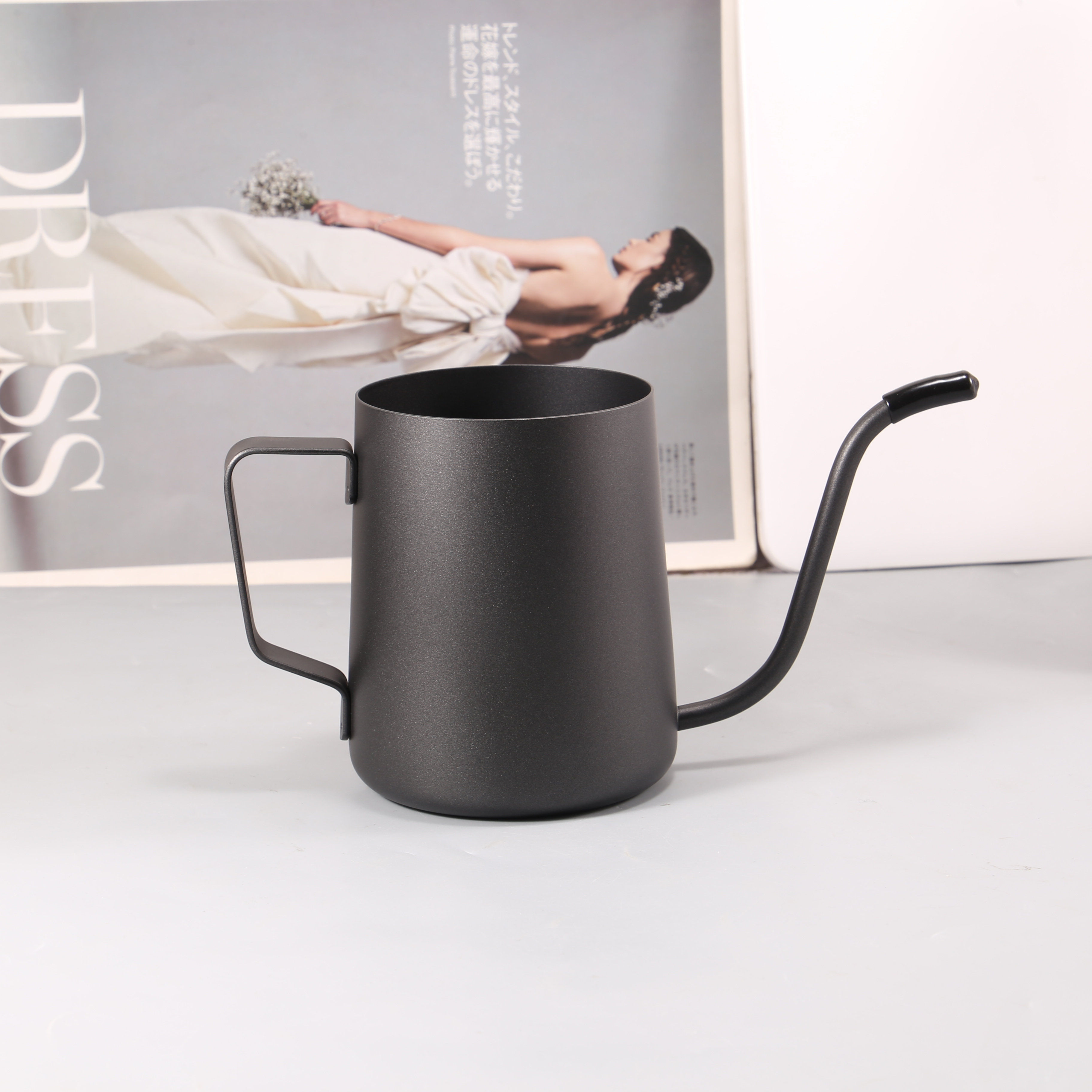 A sleek black gooseneck kettle and a high-quality 304 stainless steel milk cup from the Coffee House Essential set are placed on a light gray surface. In the background, an open magazine showcases a woman in a white dress holding a bouquet, leaning against an immaculate white wall.