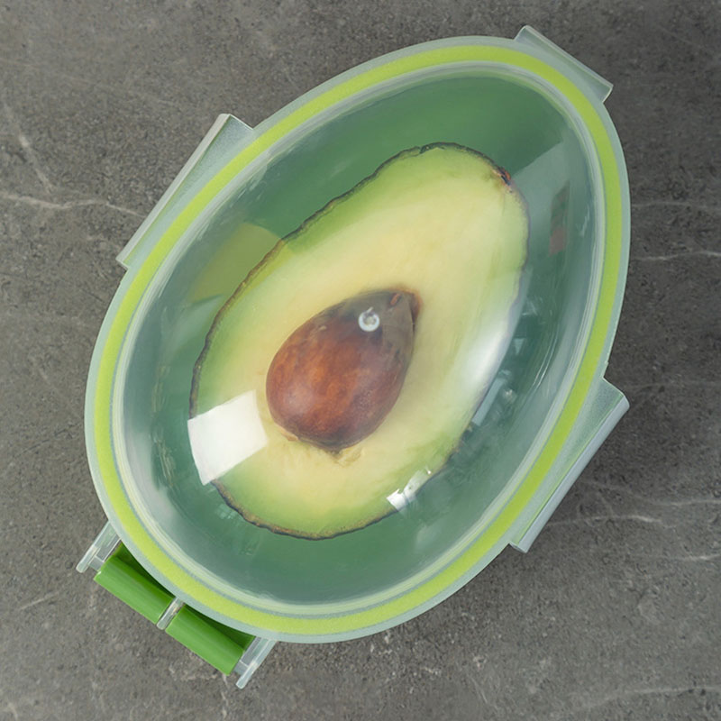 A halved avocado with its seed is stored in an Avocado Crisper Box Airtight, featuring a transparent lid, placed on a gray surface.