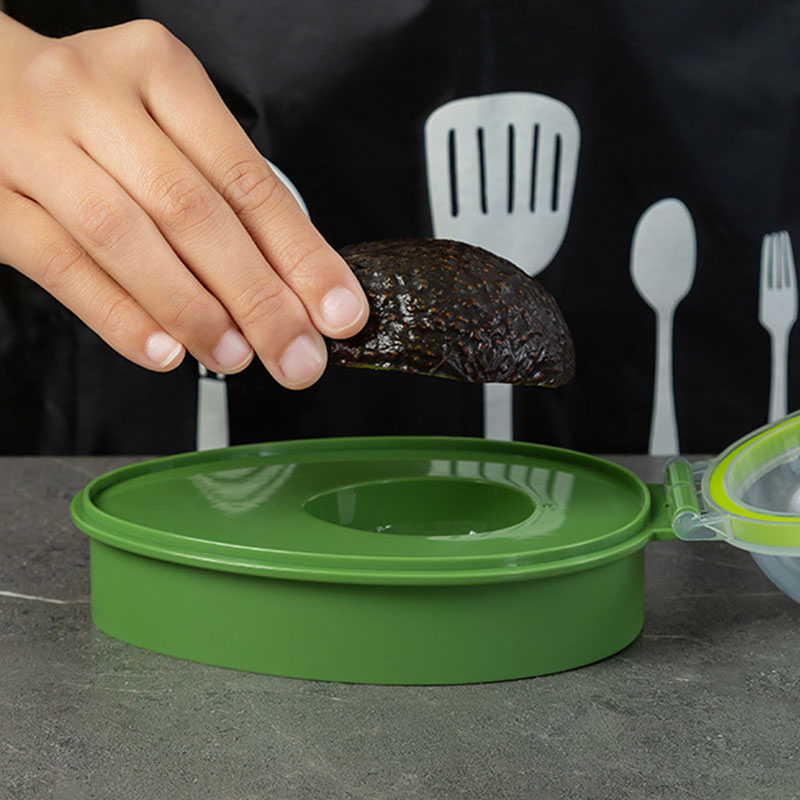 A hand places a halved avocado into the Avocado Crisper Box Airtight, also known as the Fresh Avocado Keeper, which is designed for optimal avocado storage. The container rests on a gray counter as a person in the background sports a dark apron featuring utensil designs.