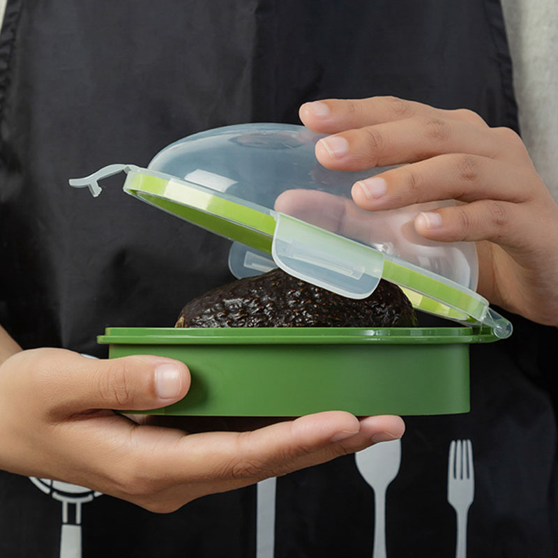 A person holds an Avocado Crisper Box Airtight, designed to preserve avocados, featuring a hinged transparent lid and revealing a portion of an avocado inside. The person is wearing a dark apron decorated with utensil patterns.