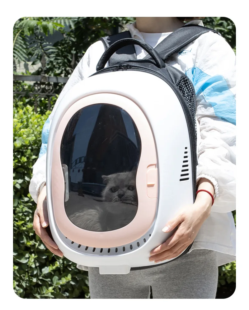 Una persona que sostiene un transportador para gatos. El transportín tiene una ventana transparente a través de la cual se puede ver un gato en su interior. La persona lleva una mochila y una chaqueta, y se vislumbra un atisbo de vegetación en el fondo, lo que sugiere un entorno al aire libre.