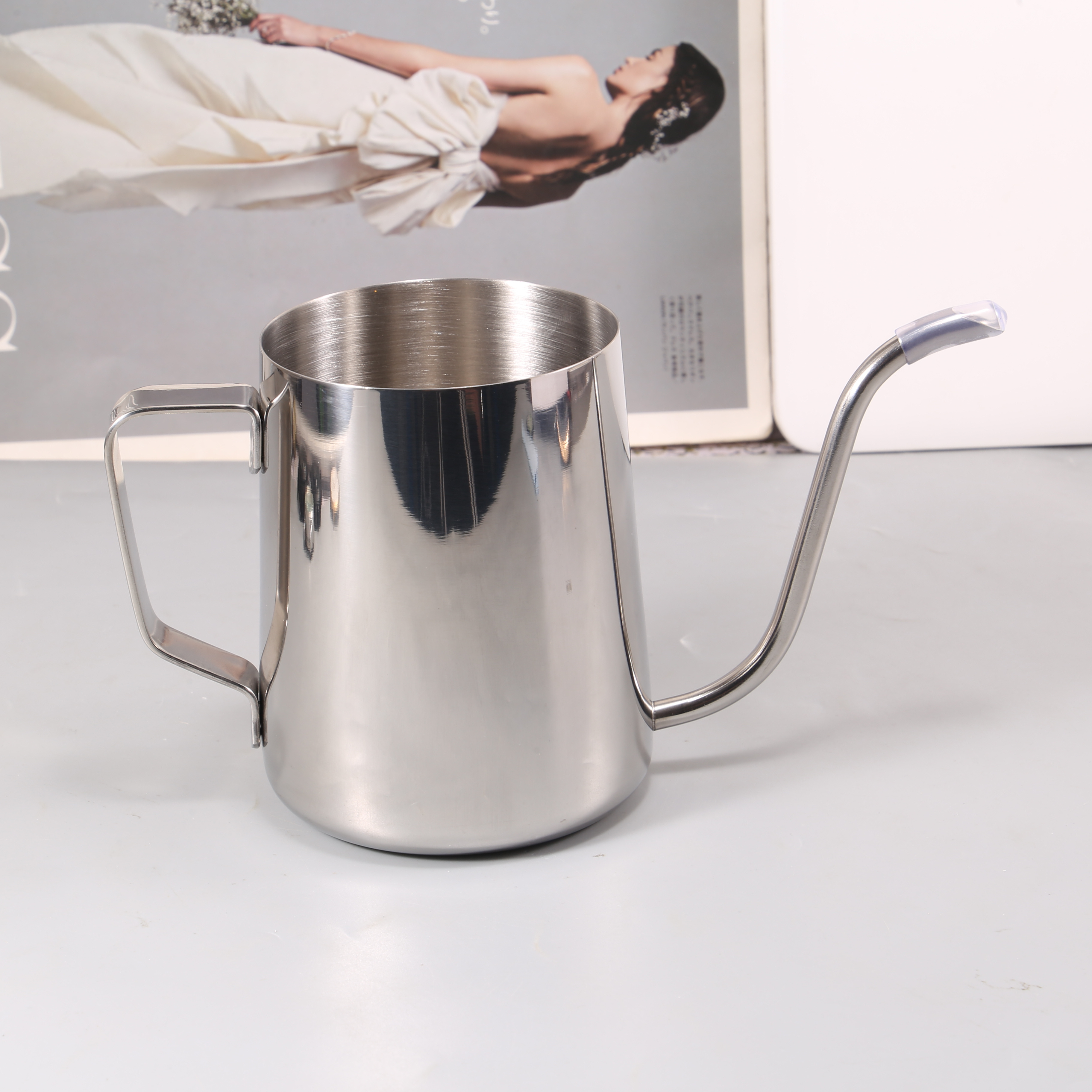 A sleek 304 Stainless Steel Milk Cup from the Coffee House Essential High-Quality Coffee Set is elegantly positioned on a light surface, while in the background, a magazine showcases a woman wearing a white dress.