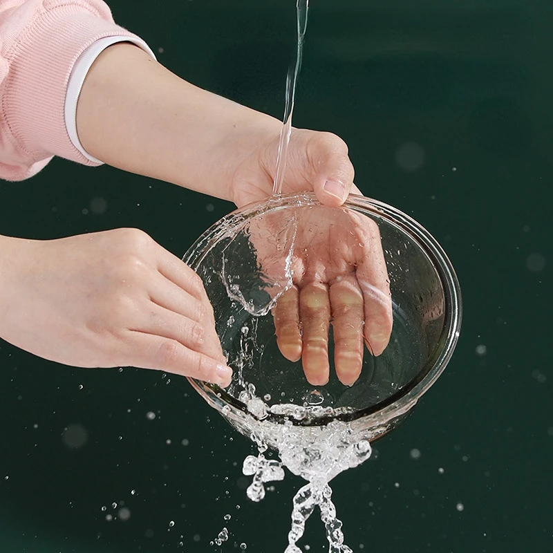 Las manos de una persona vertiendo agua en un recipiente de vidrio transparente. El agua se vierte desde una altura, creando un chorro de agua que es visible cuando cae en el recipiente. El fondo es de un color sólido, lo que proporciona un claro contraste con la acción que tiene lugar.