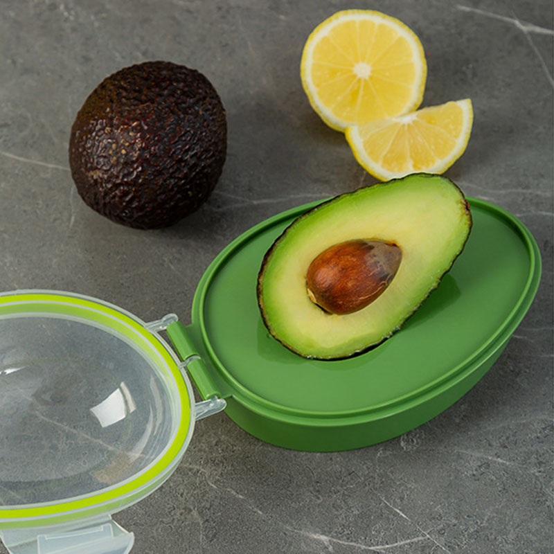 A sliced avocado is nestled in the Avocado Crisper Box Airtight, a storage container with a transparent lid, on a gray marble surface. Nearby, a whole avocado and two lemon halves add contrast, completing this elegant presentation.